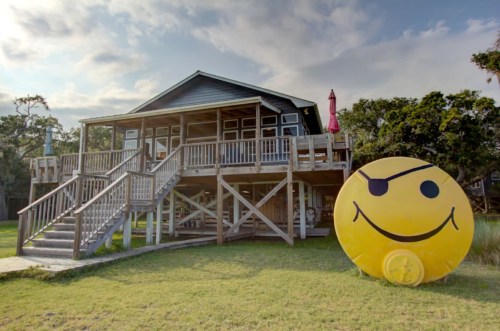 A wooden house on stilts with a large yellow smiley face sculpture in the foreground, set against a cloudy sky.