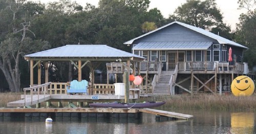 A waterfront house with a dock, kayak, and a smiling sun decoration, surrounded by trees and calm water.