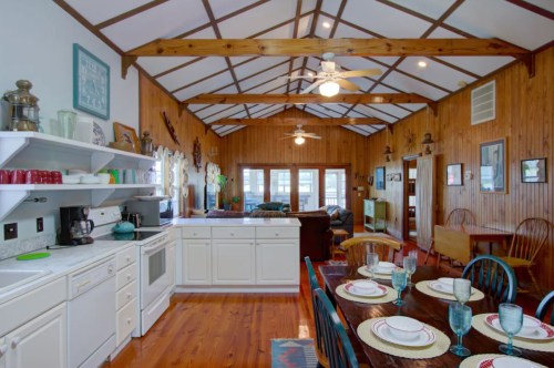 Bright, rustic kitchen with wooden beams, white cabinets, and a dining table set for a meal. Cozy living area in the background.