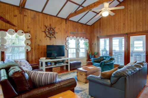 Cozy living room with wooden walls, leather sofas, a TV, and large windows, featuring nautical decor and a ceiling fan.
