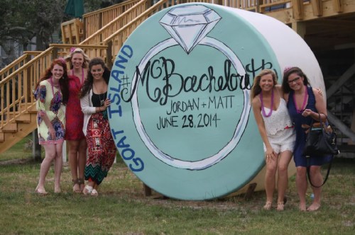 A group of six women poses in front of a large painted sign for a bachelorette party on Goat Island.