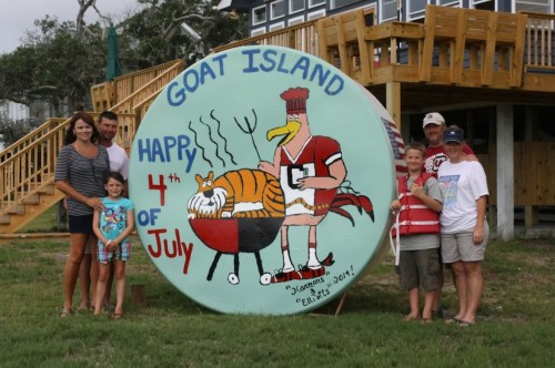 A family poses in front of a large painted sign that reads "Goat Island Happy 4th of July" with a cartoon character grilling.
