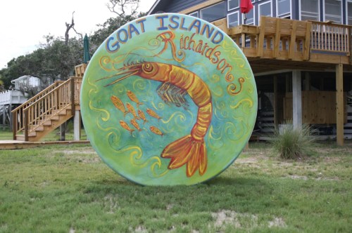 Colorful sign featuring a shrimp and fish, labeled "Richardson's Goat Island," set in a grassy area near a building.