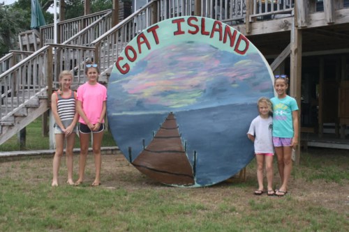Four children stand beside a large painted sign that reads "Goat Island," with a colorful sky and dock design.