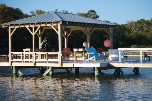 A wooden gazebo on a dock over calm water, featuring chairs and decorative floats, surrounded by trees.