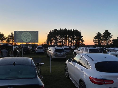 A drive-in movie scene at sunset, with cars parked in front of a large screen and trees in the background.