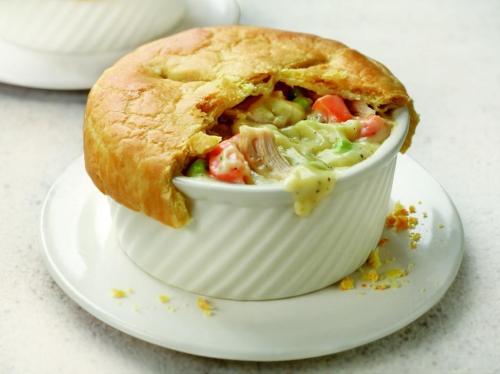 A creamy chicken pot pie in a white ramekin, topped with a golden, flaky crust and filled with colorful vegetables.