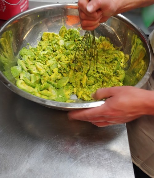 A person whisking a green mixture in a large metal bowl, with visible avocado pieces and a vibrant color.