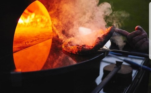 A hand lifts a freshly baked pizza from a wood-fired oven, with smoke and glowing embers visible in the background.