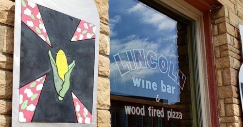 A colorful sign featuring corn hangs beside a window displaying "Lincoln Wine Bar & Wood Fired Pizza."