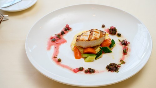 Grilled fish fillet on a bed of vegetables, garnished with sauce and capers, served on a white plate.