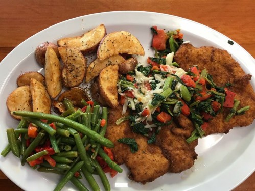 A plate of fried chicken with sautéed vegetables, seasoned potatoes, and green beans on a wooden table.