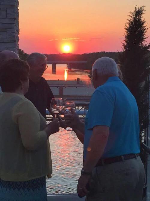 A group of people toasting with glasses at sunset by a river, with vibrant colors reflecting on the water.