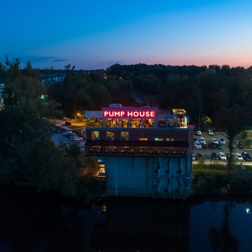 Aerial view of the Pump House restaurant at dusk, illuminated sign, surrounded by trees and parked cars by the water.