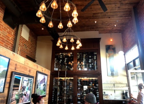 Interior of a cozy restaurant with wooden beams, a chandelier, and wine bottles displayed in a cabinet.