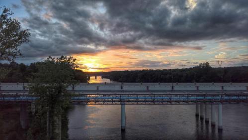 Sunset over a river, with clouds in the sky and a bridge crossing the water, surrounded by trees and hills.