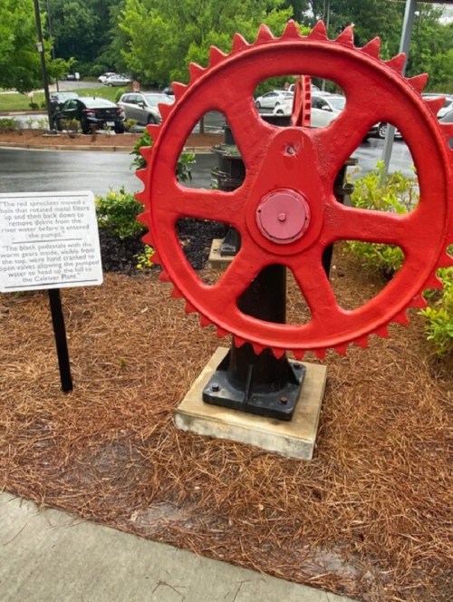 A large red gear sculpture stands beside a sign in a landscaped area, with cars and trees in the background.