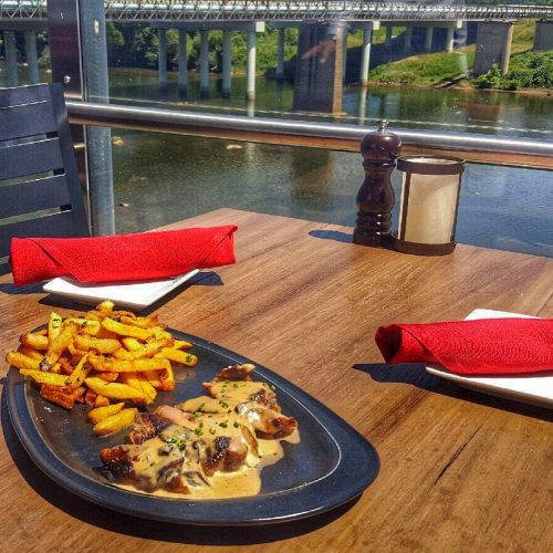 A plate of chicken in sauce with fries, set on a table overlooking a river and bridge on a sunny day.