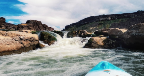 A kayak approaches rocky rapids with rushing water under a cloudy sky.