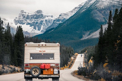 A vintage RV drives down a winding road surrounded by mountains and evergreen trees under a cloudy sky.