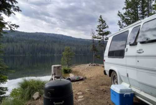 A serene lakeside scene featuring a van, cooler, and grill, surrounded by trees and mountains under a cloudy sky.