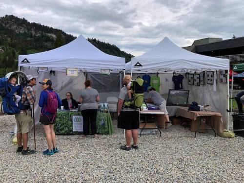 Two white tents at an outdoor event, with people browsing merchandise and information displays. Mountains in the background.