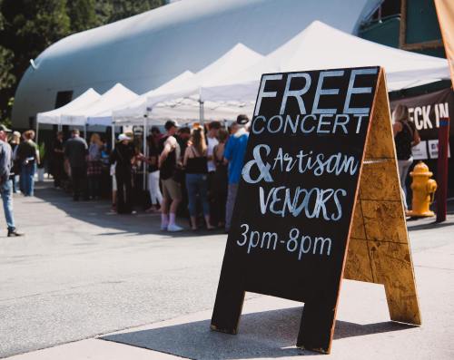 A sign advertising a free concert and artisan vendors, with people lined up under tents in the background.