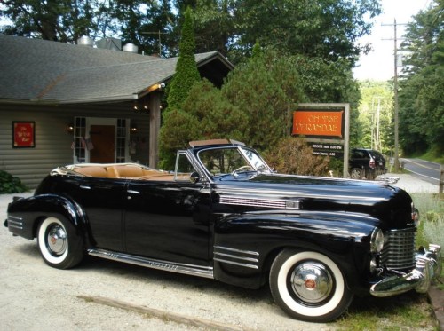 A classic black convertible car parked outside a rustic building surrounded by trees.