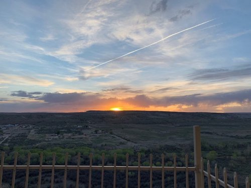 Sunset over a vast landscape with rolling hills, green trees, and a colorful sky, framed by a fence in the foreground.