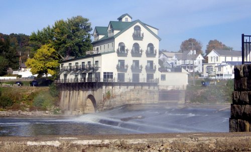 A riverside building with balconies overlooks a waterfall, surrounded by trees and houses in the background.