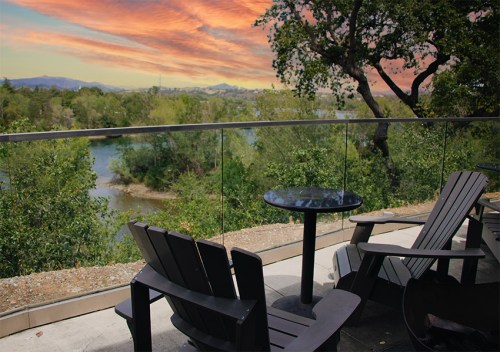 A serene view from a balcony with two chairs and a table overlooking a river and lush greenery at sunset.