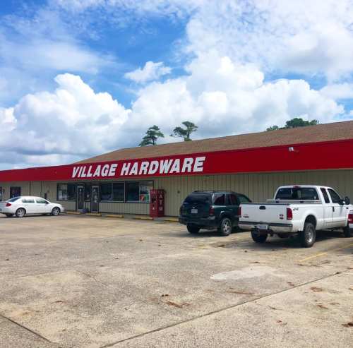 A hardware store with a red roof, featuring parked cars in front and a blue sky with clouds above.