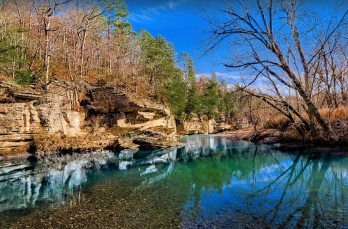 A serene river flows through a rocky landscape, surrounded by trees and reflecting the clear blue sky.