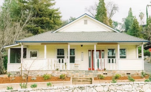 A charming yellow house with a porch, surrounded by trees and landscaped with small plants and gravel.