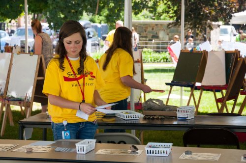 Two volunteers in yellow shirts work at a table with art supplies under a tent at an outdoor event.