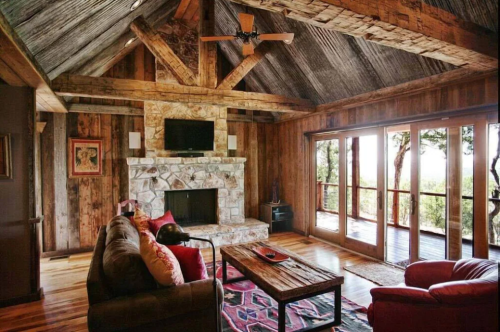 Cozy rustic living room with wooden beams, stone fireplace, and large windows overlooking nature.