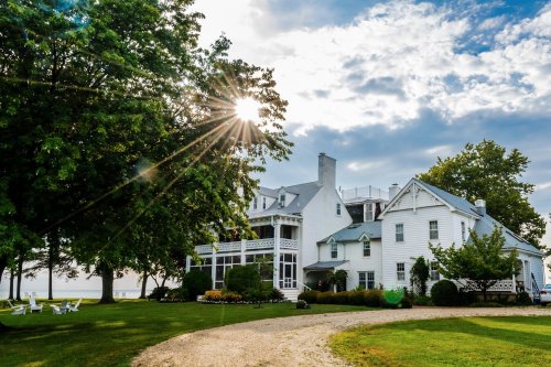 A large white house with a wraparound porch, surrounded by trees and a grassy lawn, under a partly cloudy sky.