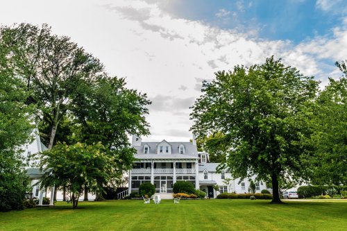 A large, elegant house surrounded by lush green trees and a manicured lawn under a partly cloudy sky.