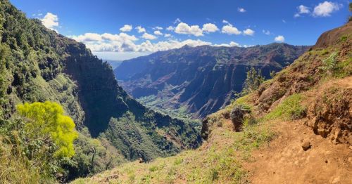 A panoramic view of a lush green valley surrounded by steep cliffs under a bright blue sky with scattered clouds.