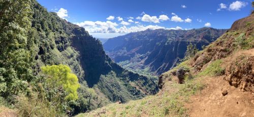 A panoramic view of a lush green valley surrounded by steep cliffs under a bright blue sky with scattered clouds.