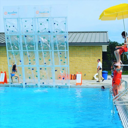 A pool scene featuring a climbing wall, children playing, and lifeguards on duty under a yellow umbrella.