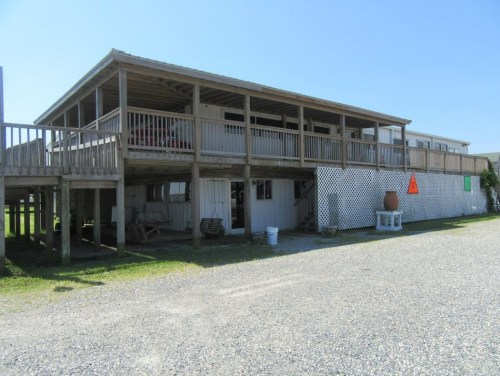 A wooden building on stilts with a large deck, surrounded by gravel and grass under a clear blue sky.