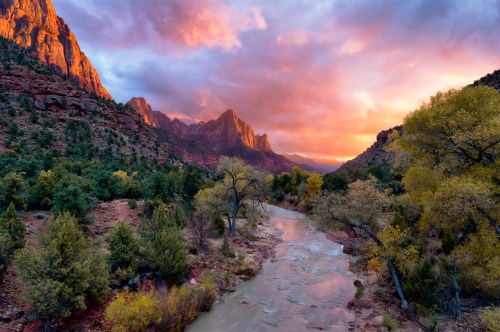 A scenic view of a river winding through a canyon, surrounded by colorful mountains and a dramatic sunset sky.