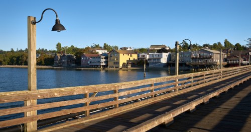 A wooden pier extends over calm water, with colorful houses lining the shore under a clear blue sky.