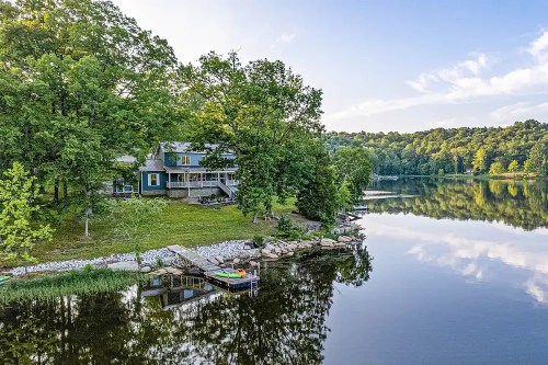 A serene lakeside view featuring a house surrounded by trees, with a dock and calm water reflecting the sky.