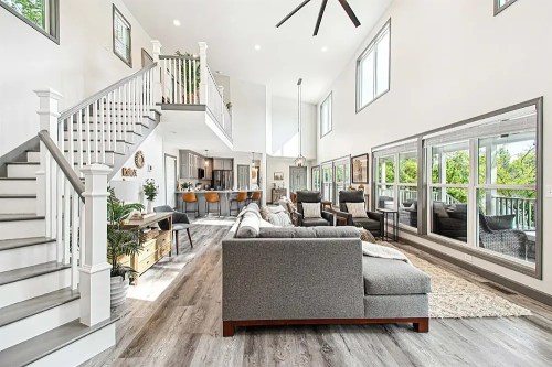 Bright, modern living room with large windows, gray sectional sofa, and open staircase leading to a second floor.
