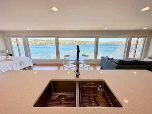 A modern kitchen sink with a view of a lake and seating area through large windows. Bright and airy interior.