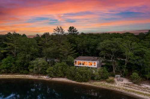 A serene lakeside house surrounded by trees, with a vibrant sunset sky reflecting on the water.