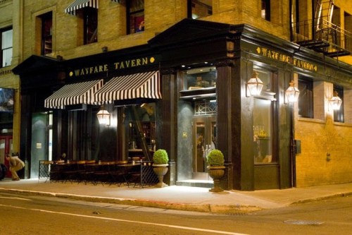 Exterior of Wayfare Tavern at night, featuring black walls, striped awnings, and warm lighting.