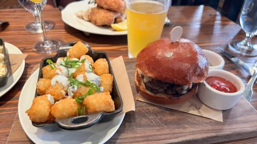 A plate with crispy tater tots topped with green onions and a burger on a brioche bun, served with ketchup and a drink.